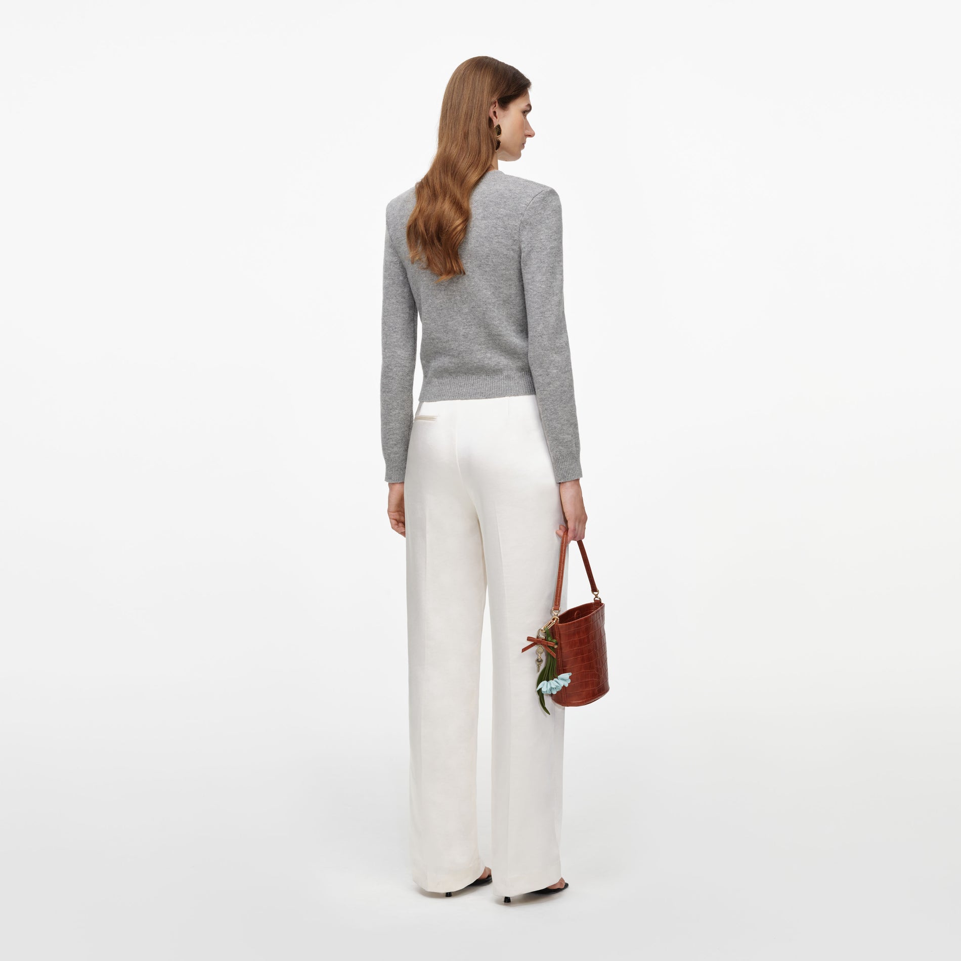 Woman facing away wearing a grey long-sleeved top, white wide-leg trousers, and holding a brown handbag