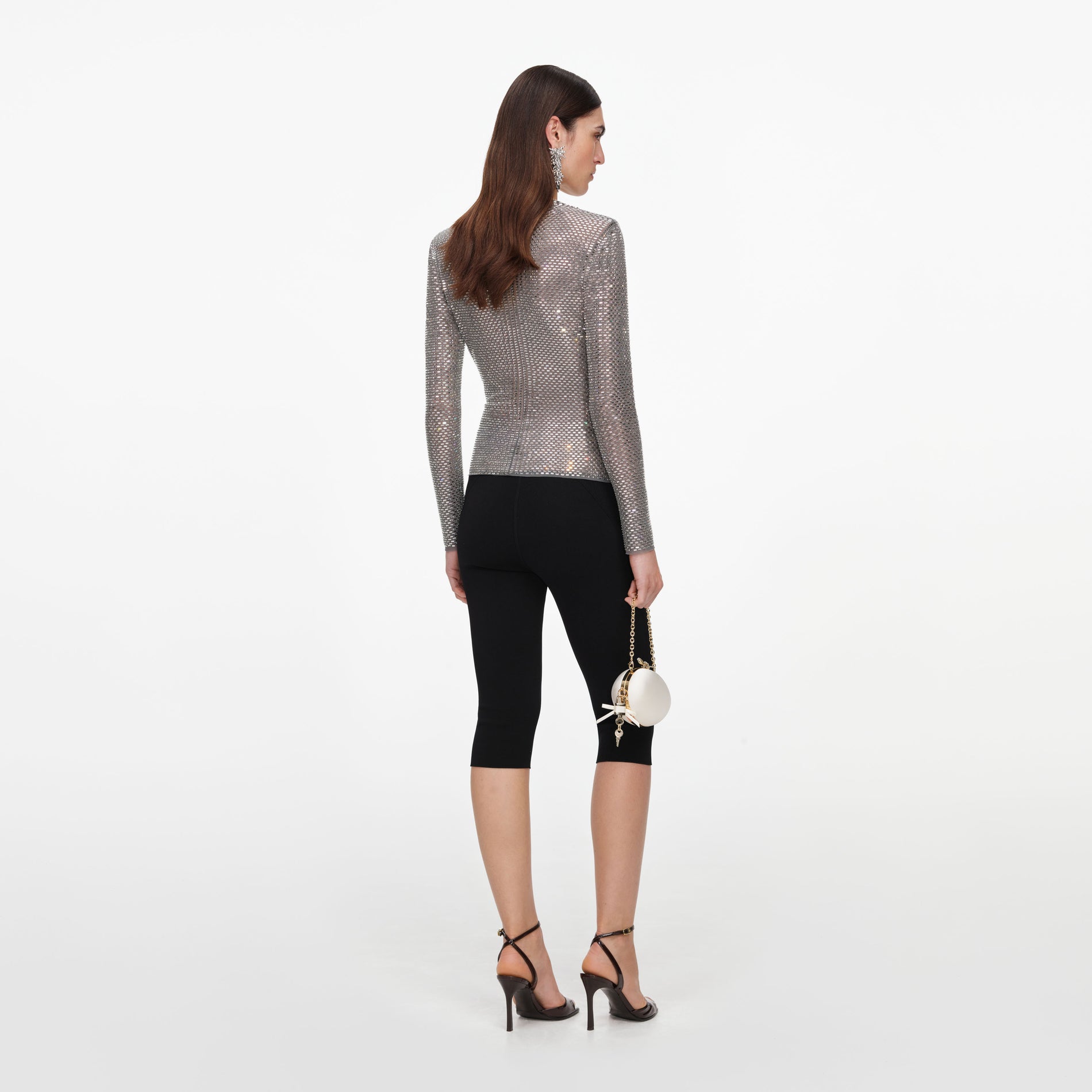 Woman in a silver long-sleeved top, black capri pants, black heels, and holding a white spherical purse